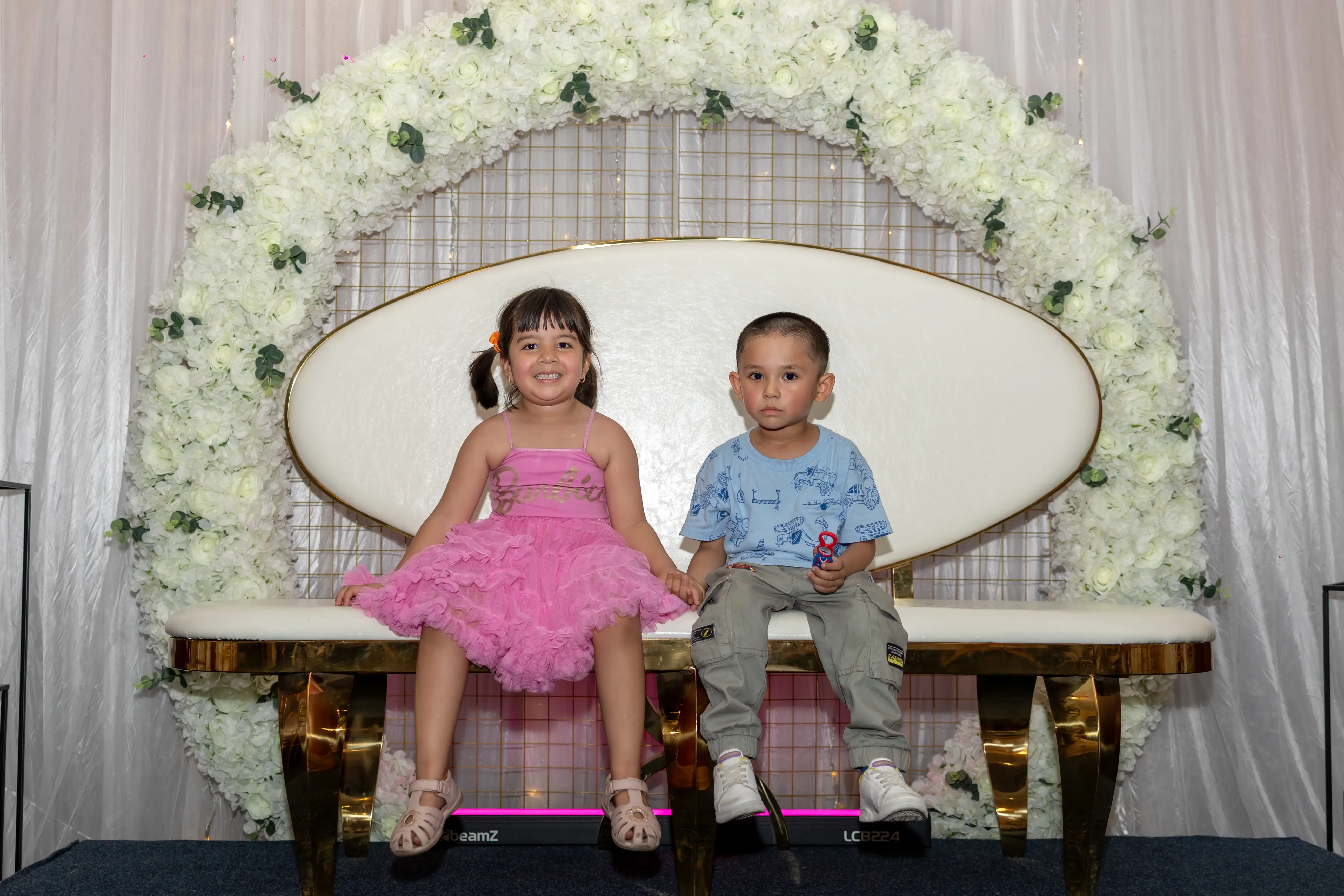 Two young children sitting side by side on a seating arrangement. The girl on the left is wearing a pink dress, and the boy on the right is in formal attire. They are enjoying a function with white flowers, greenery, and an arch-shaped decoration at Third Eye Windsor, 515 George St, South Windsor NSW 2756.