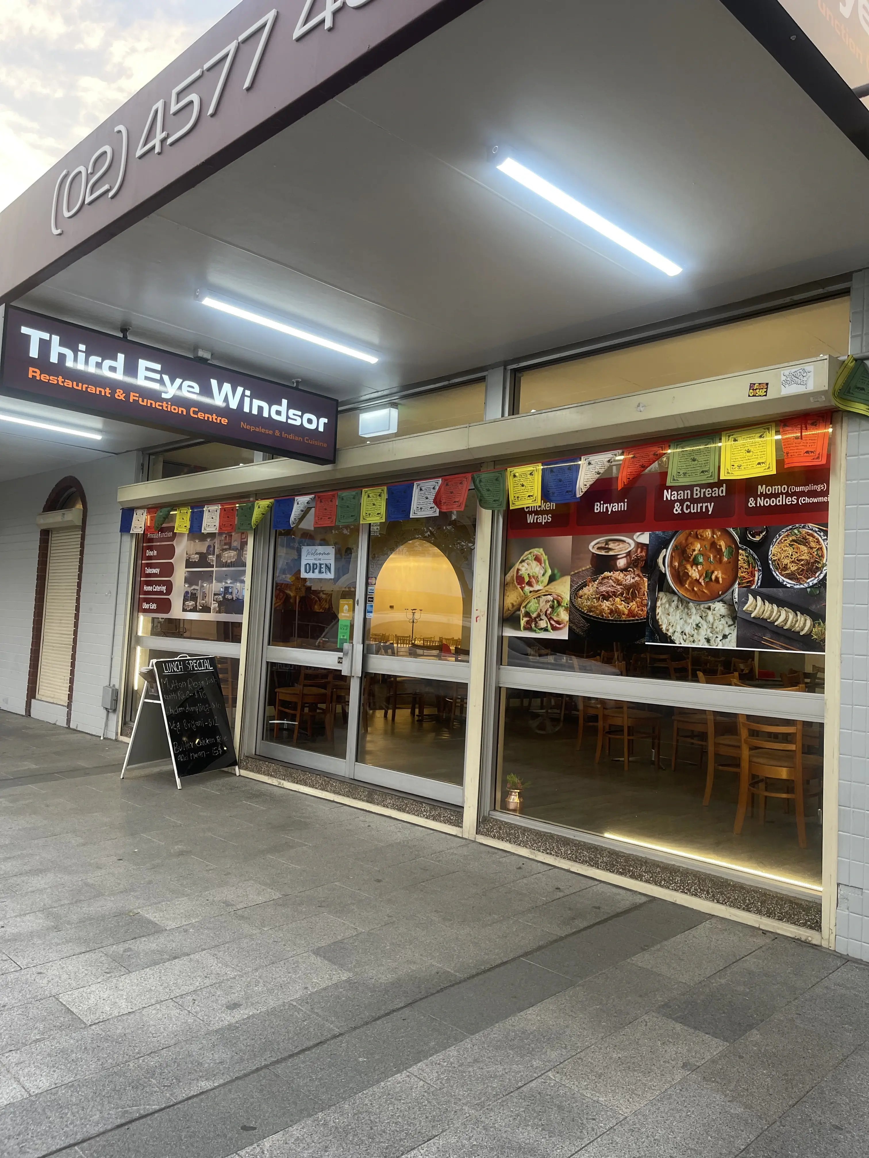 Street view of Third Eye Windsor Restaurant & Function Centre, showcasing its glass-front entrance with colorful Tibetan prayer flags. A sign displays the restaurant's name. The window features large food images promoting biryani, naan bread, and curry. A board near the entrance indicates that the restaurant is open. Interior wooden chairs and tables are visible through the windows  at 515 George St, South Windsor NSW 2756.