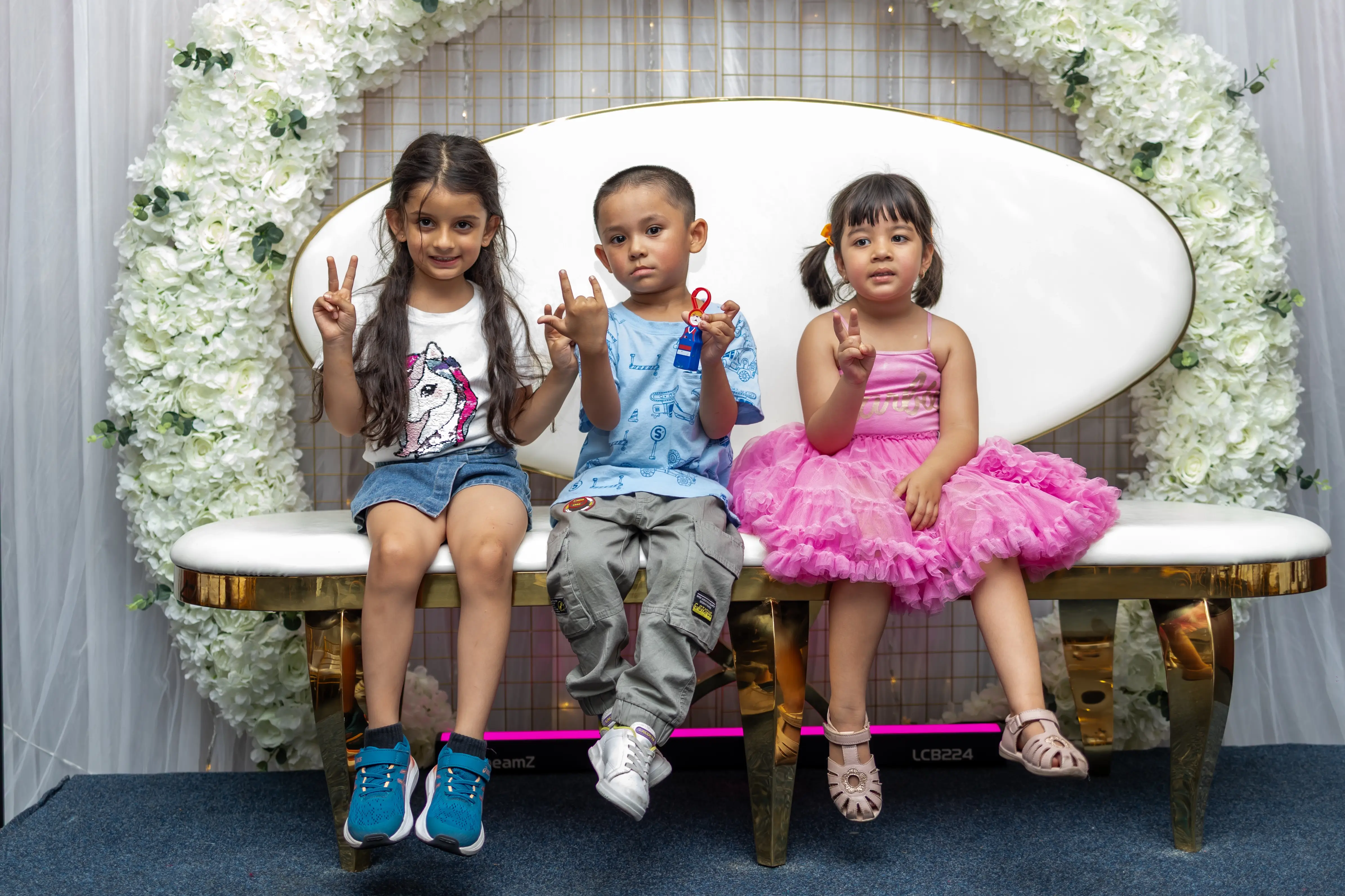 Three young children sitting side by side on a seating arrangement at Third Eye Windsor, 515 George St, South Windsor NSW 2756. The girl on the left is wearing a white and blue dress, the boy in the middle is in formal attire, and the girl on the right is dressed in pink. They are enjoying a function, surrounded by white flowers, greenery, and an arch-shaped decoration, and are posing for a photo.