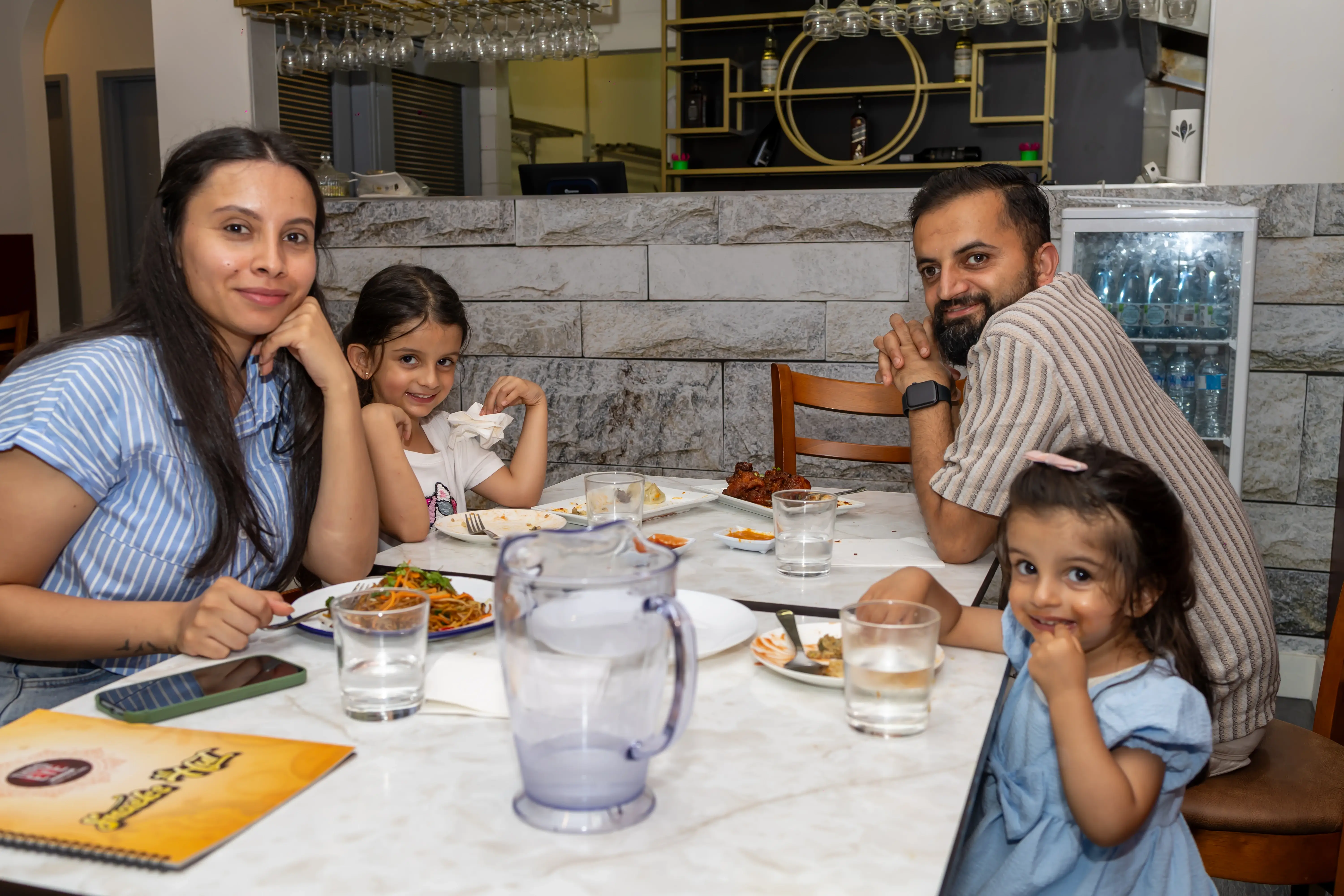 A family of four at Third Eye Windsor, with a couple and two young girls enjoying their meal. All are smiling, with the kids enjoying kid-friendly menu items while the couple has chowmein. The table is full of food, and the baby looks especially happy enjoying their meal at 515 George St, South Windsor NSW 2756.