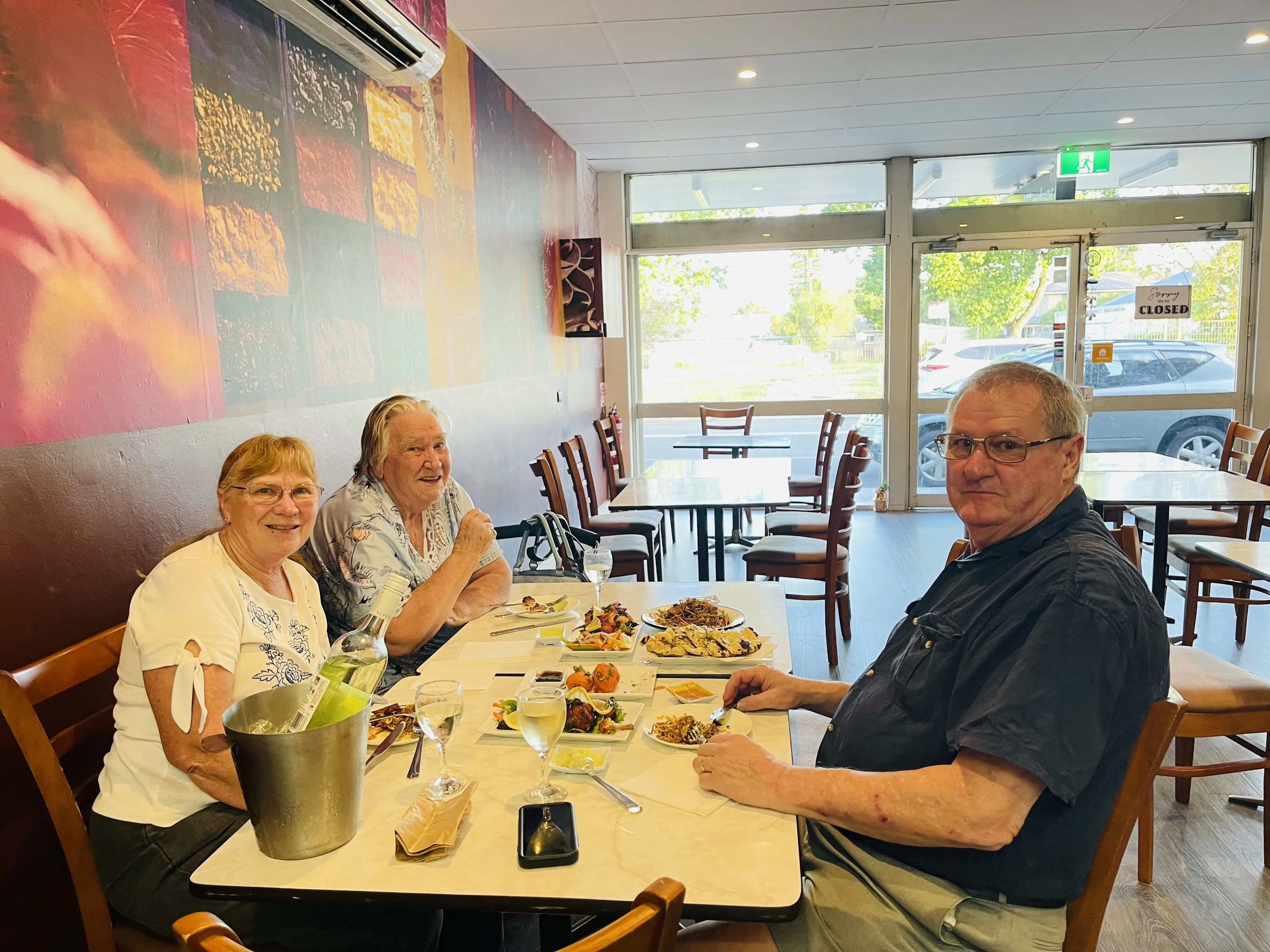 Three people seated at a dining table in a restaurant, enjoying a meal together. Two women sit on one side of the table, with a man in a dark blue shirt across from them. The table is filled with various Indian and Nepali dishes at Third Eye Windsor, 515 George St, South Windsor NSW 2756.