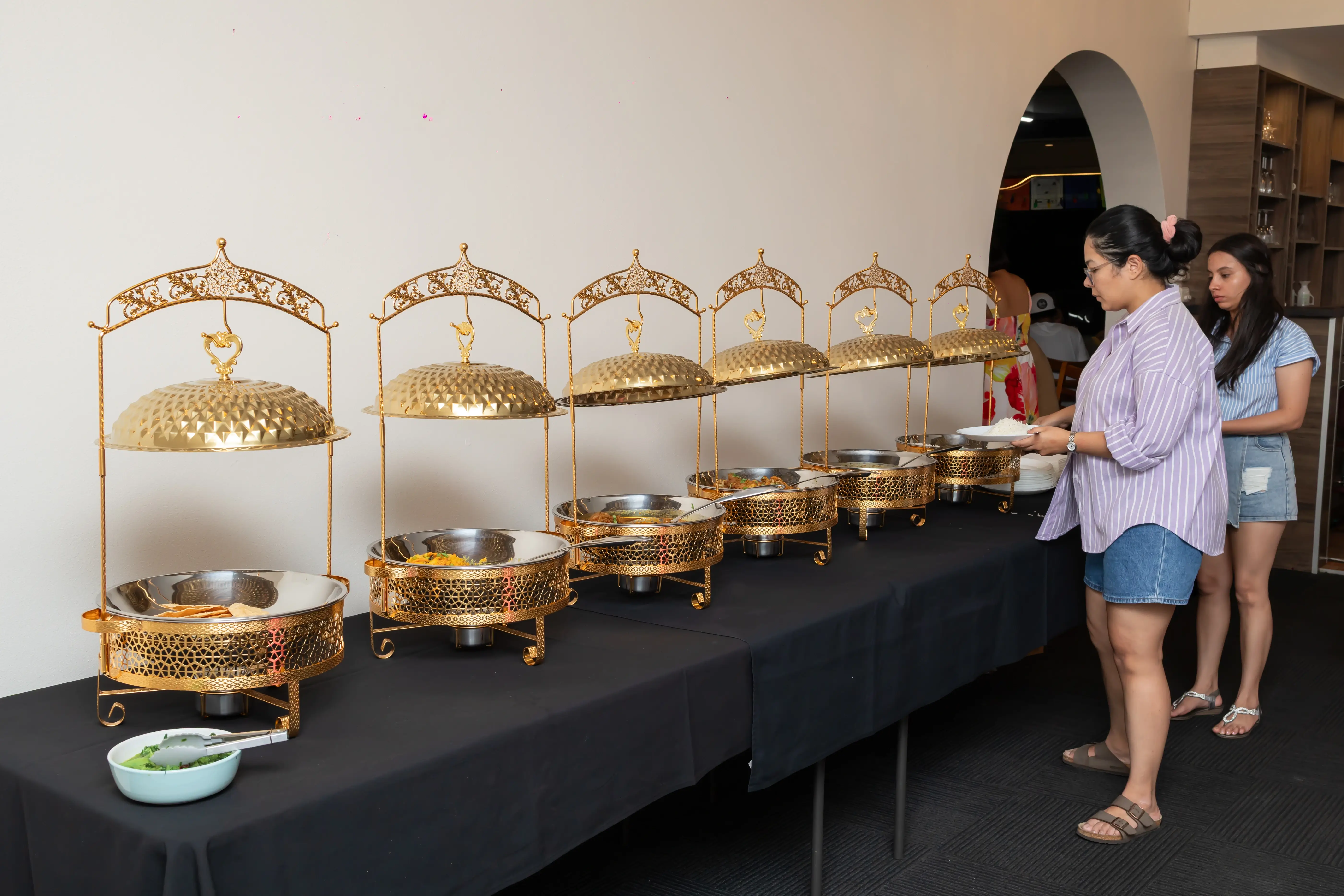 A buffet setup covered with black tablecloths, featuring chafing dishes on decorative gold-colored stands with elaborately designed domed lids. The arrangement is for a catered event. On the right side of the image, two people are serving themselves from the buffet at Third Eye Windsor, 515 George St, South Windsor NSW 2756.
