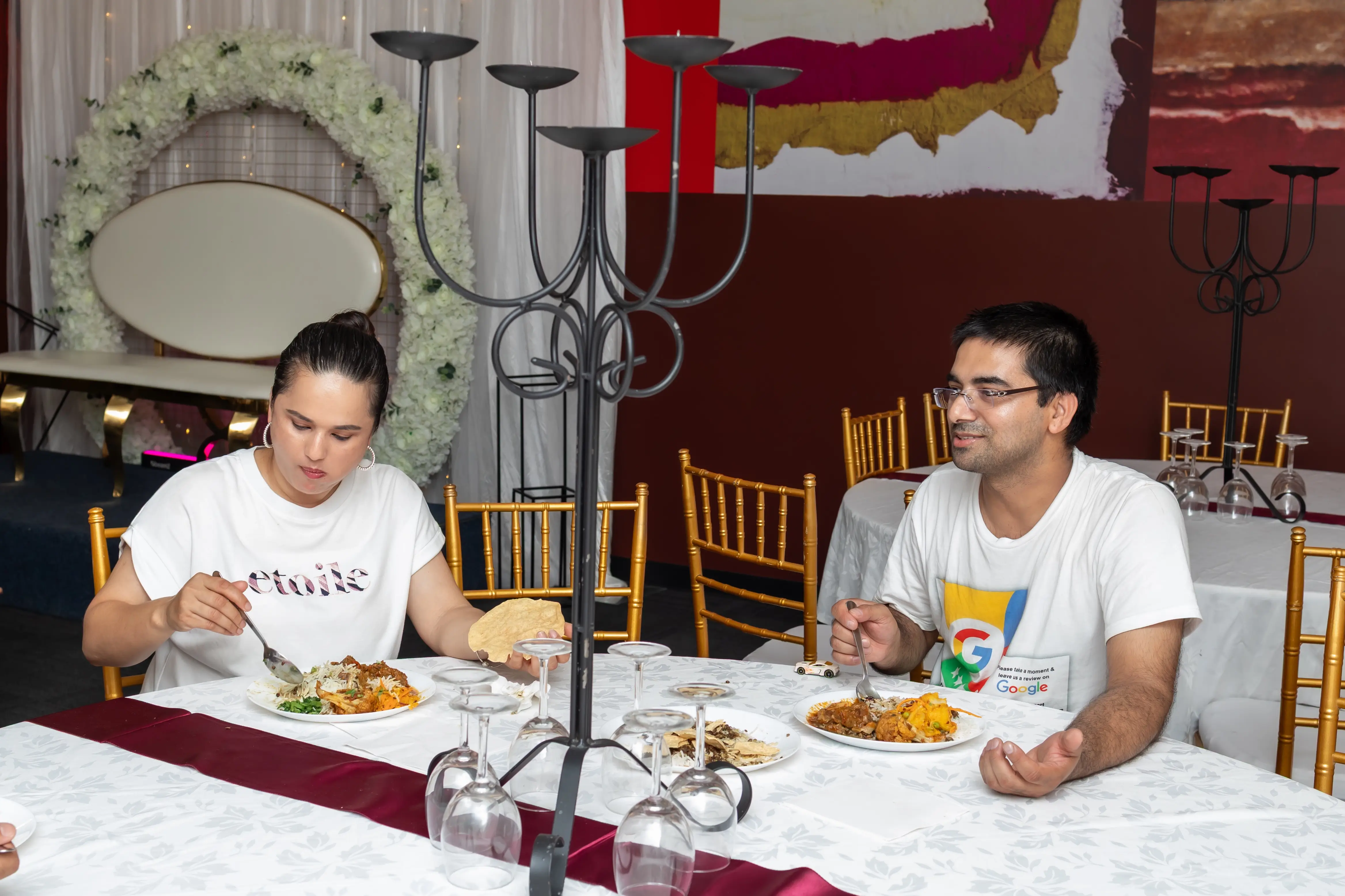 A couple dining at a formally set table in an event venue. They are seated at a table with a white tablecloth, and both are wearing white t-shirts and enjoying a meal together. A decorative candelabra-style centerpiece is placed in the center of the table. Behind them is a circular white floral decoration and gold Chiavari chairs at Third Eye Windsor, 515 George St, South Windsor NSW 2756.