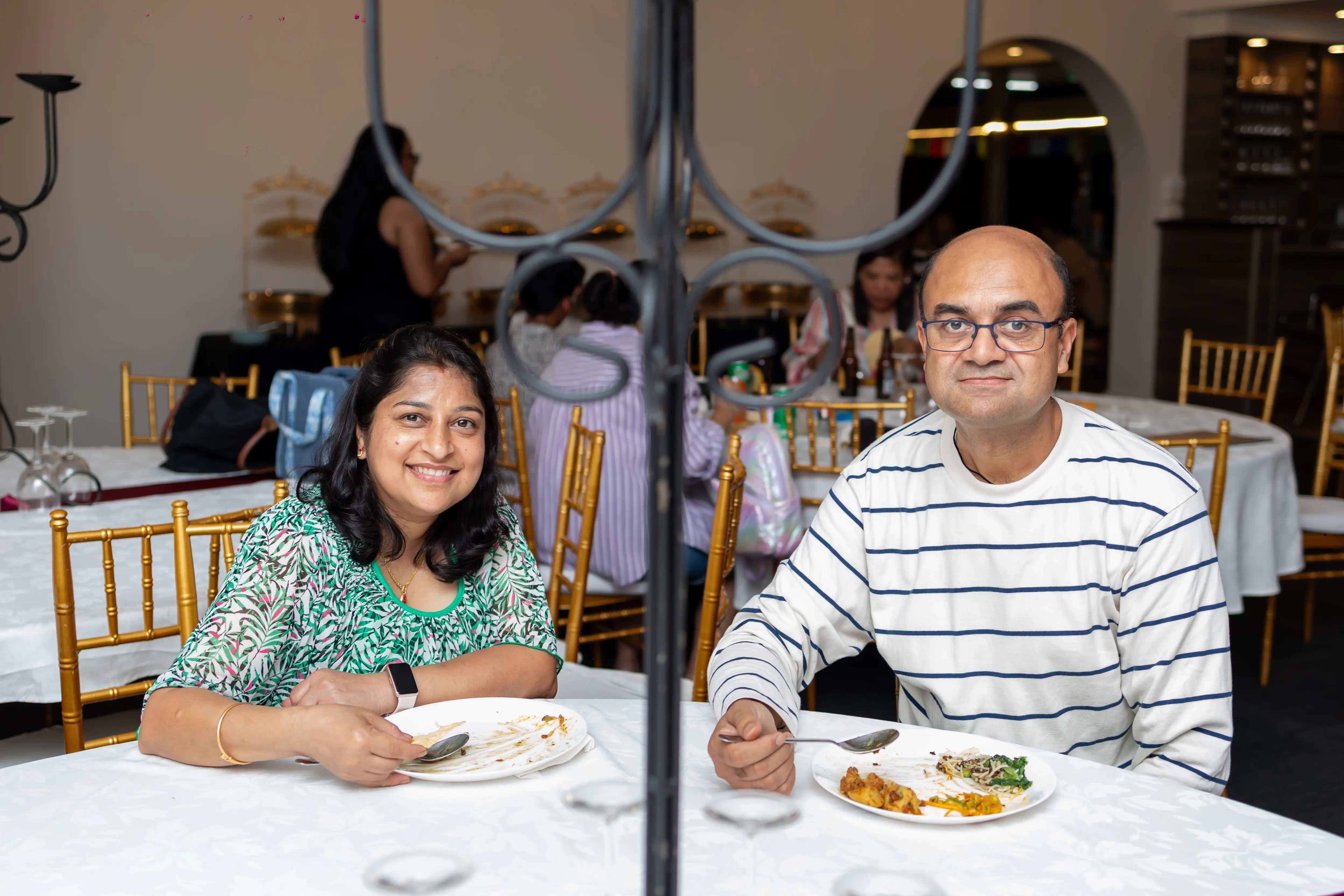 A couple dining at a formally set table in an event venue at Third Eye Windsor, 515 George St, South Windsor NSW 2756. They are dressed in semi-formal attire, enjoying a meal together, and smiling for the photo. The table is covered with a white tablecloth and features a decorative candelabra-style centerpiece. In the background, other guests are also dining and seated on gold Chiavari chairs.