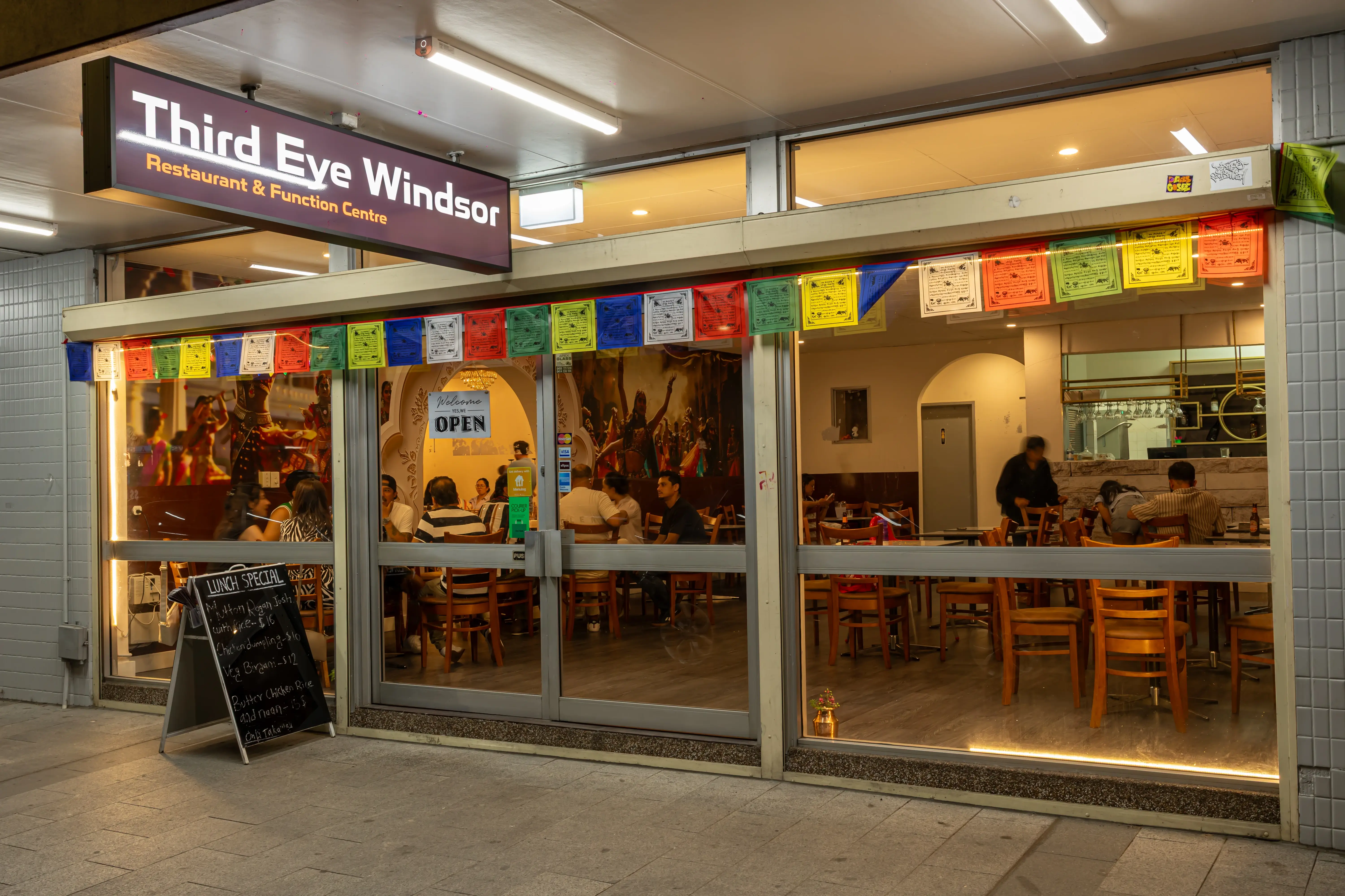 The exterior of Third Eye Windsor restaurant at night, warmly lit and inviting. The entrance features large glass windows and doors with wooden frames, and colorful Tibetan prayer flags hanging above. Diners can be seen enjoying meals inside, adding to the cozy and welcoming ambiance at 515 George St, South Windsor NSW 2756.