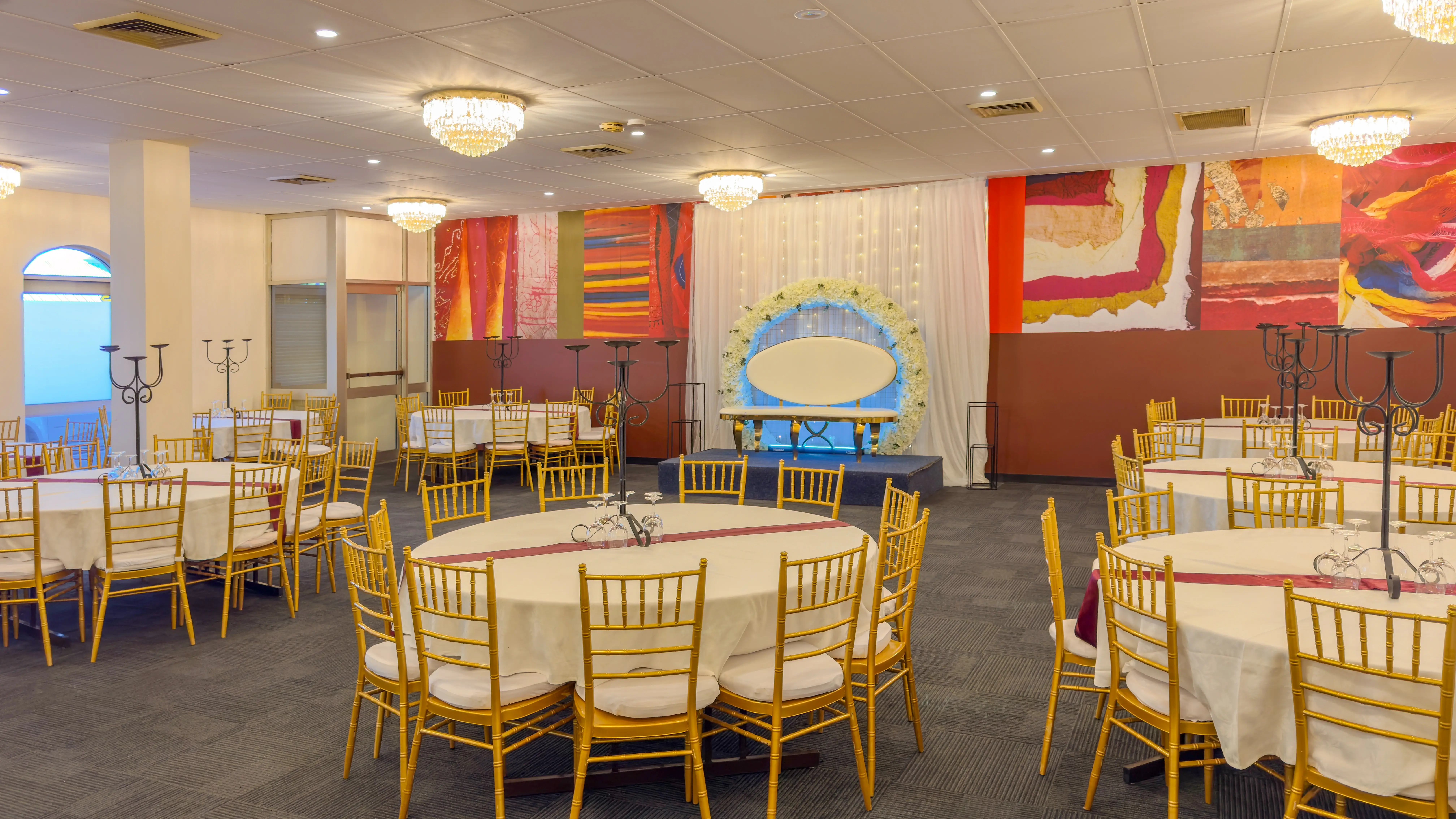 Event space arranged for a formal gathering. The room features several round tables with white tablecloths, each surrounded by elegant gold Chiavari chairs with cream-colored cushions. The tables appear to be set up for a celebration or formal dinner. In the background,