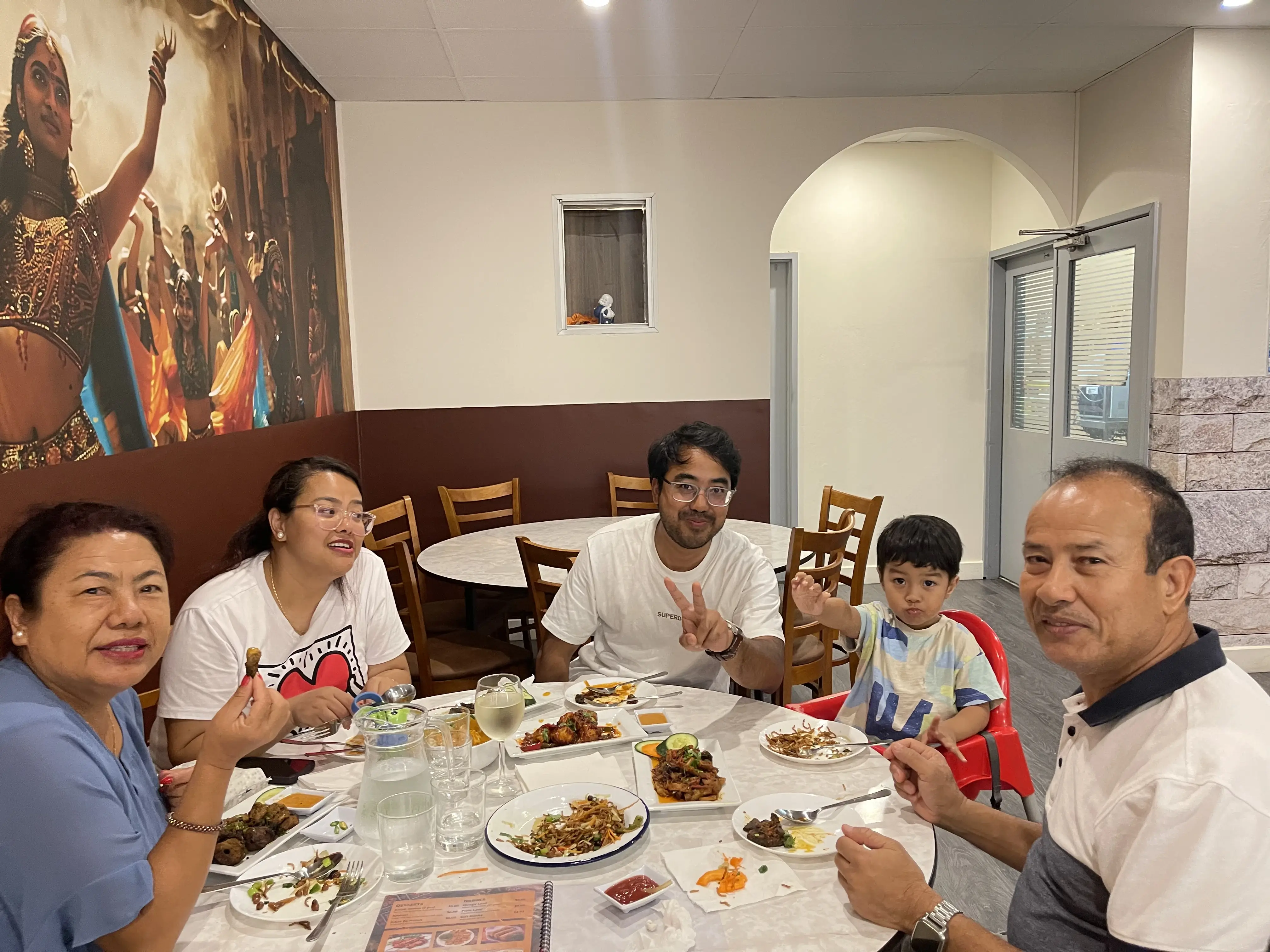 A family seated at a dining table in a restaurant, enjoying a meal together. Four adults and one young boy are present. The boy is enjoying a kids' menu, while the others are having Nepalese and Indian cuisine at Third Eye Windsor, 515 George St, South Windsor NSW 2756.