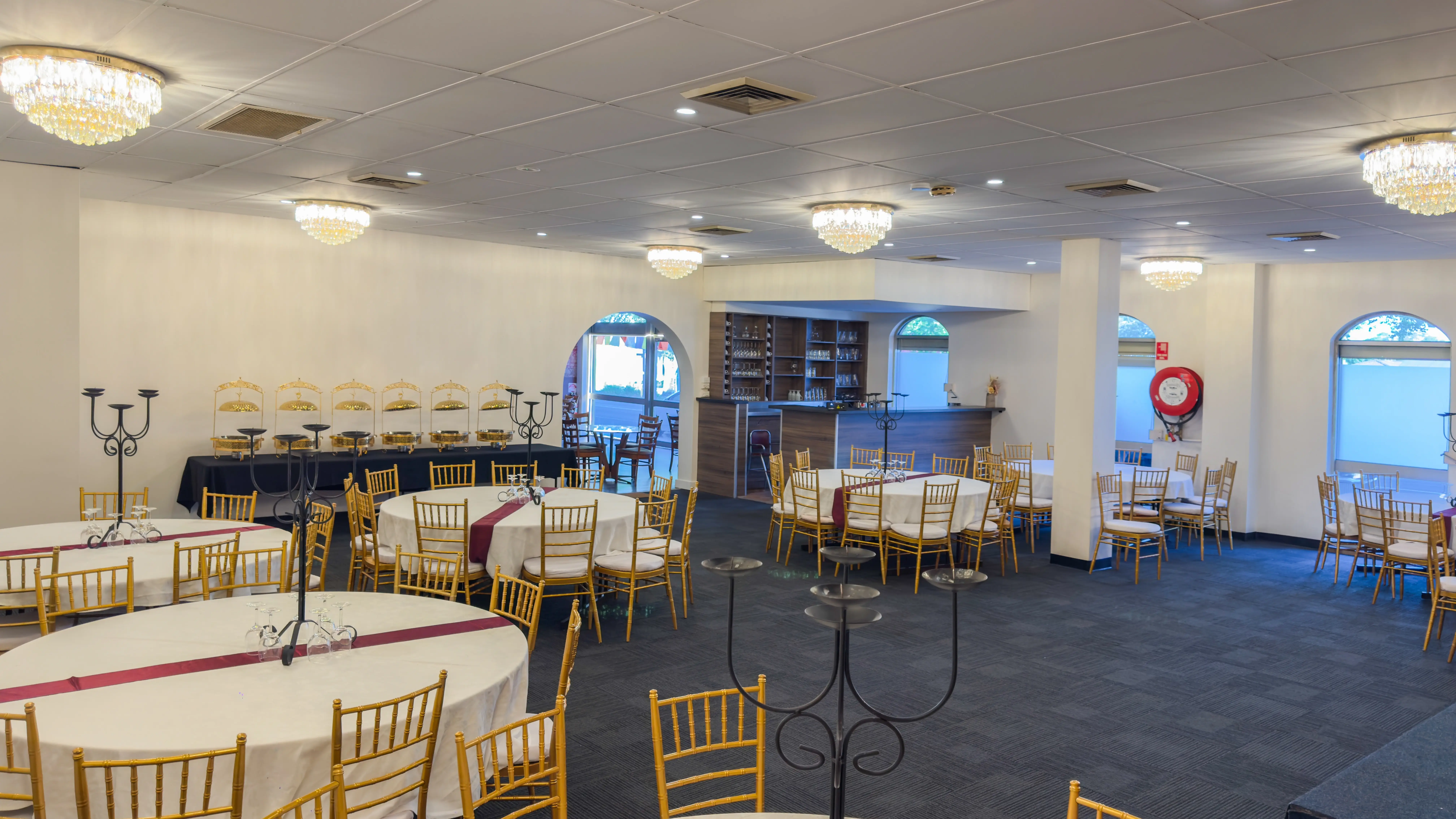 The event venue for a formal gathering at Third Eye Windsor, 515 George St, South Windsor NSW 2756. The space features multiple round tables with white tablecloths, each surrounded by gold Chiavari chairs. The room has a modern design with lighting and decorative crystal chandeliers hanging from the white ceiling.