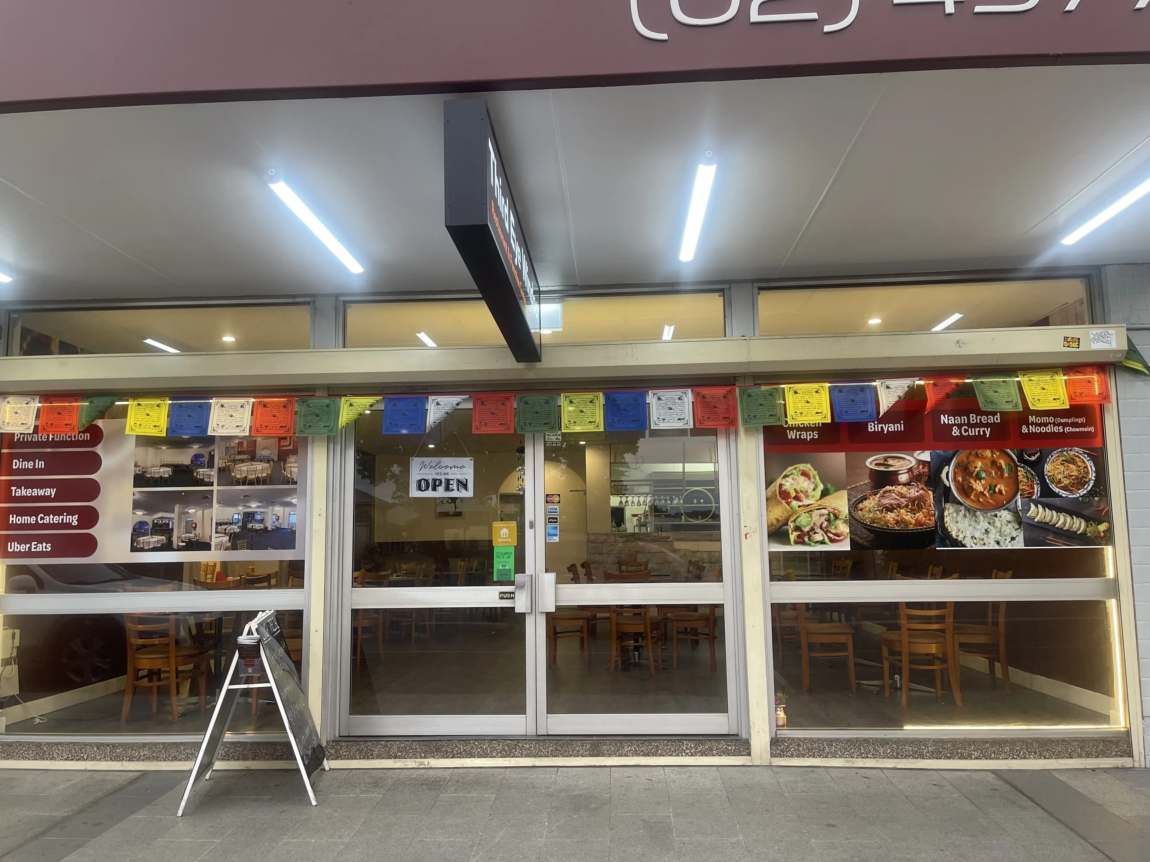 The exterior entrance of Third Eye Windsor restaurant with glass doors and windows framed in wood. Colorful Tibetan prayer flags hang above the entrance, reflecting Nepalese or Himalayan cultural influence. Food photos are displayed on the glass, showcasing various dishes from the menu at Third Eye Windsor, 515 George St, South Windsor NSW 2756.