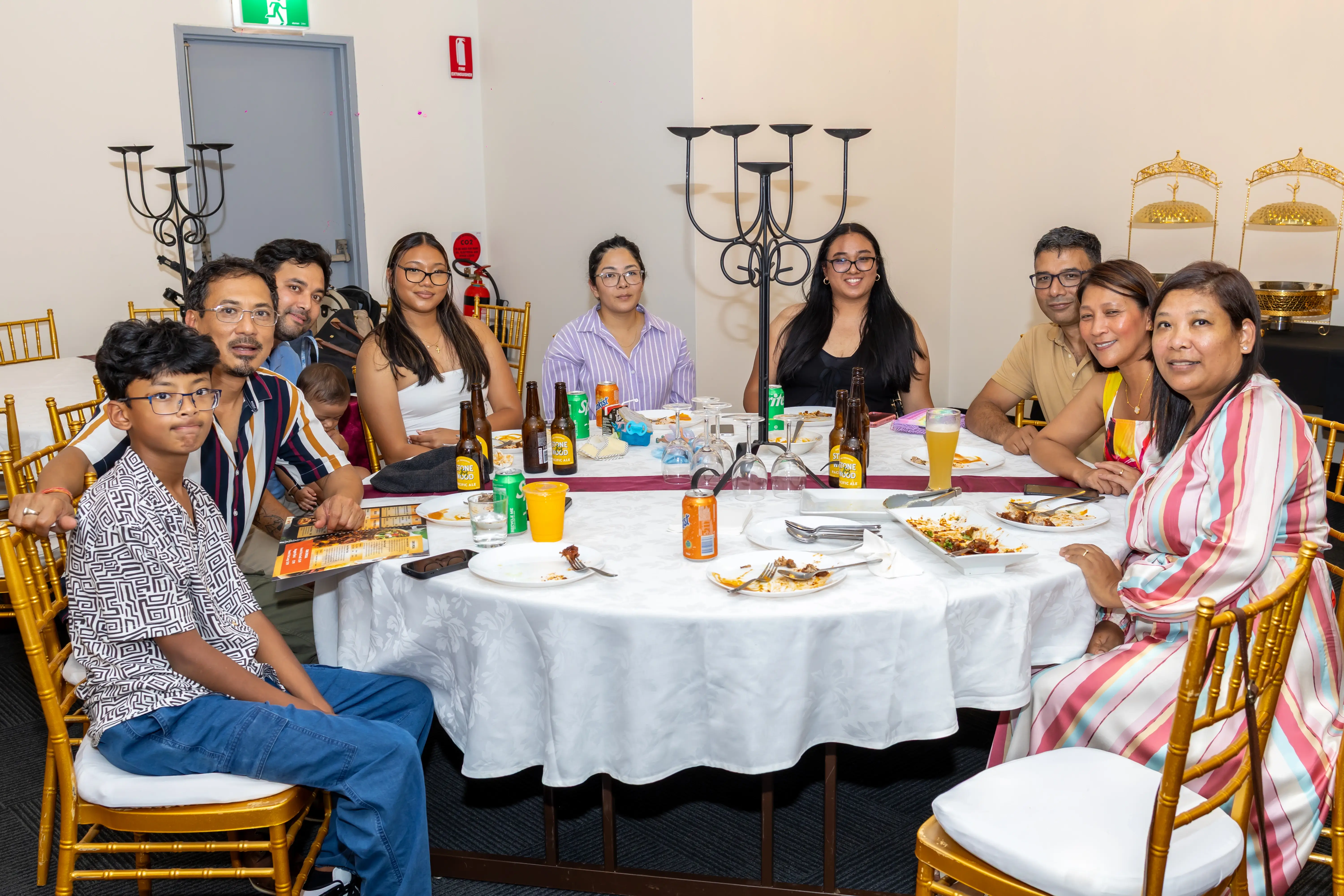 A group of family members and friends enjoying a meal together, seated around a round table covered with a white tablecloth. The table has plates of food, bottles, and colorful drinks. All are smiling at the camera, enjoying a function at Third Eye Windsor, 515 George St, South Windsor NSW 2756.