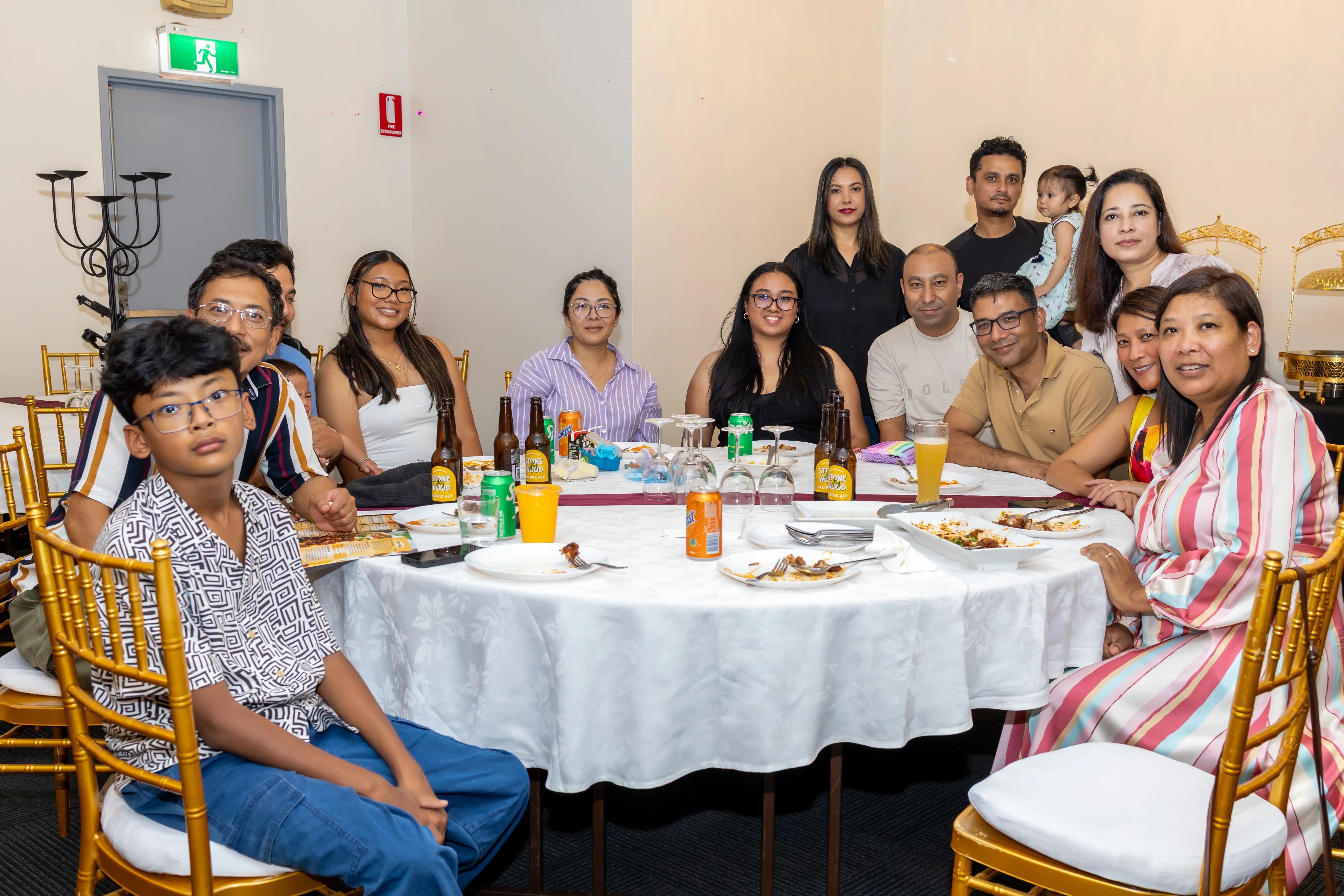 A group of family members enjoying a meal together. Some are seated and enjoying food, while others are standing around a round table covered with a white tablecloth. The table has plates of food, bottles, and colorful drinks. All are smiling at the camera, having fun at the function at Third Eye Windsor, 515 George St, South Windsor NSW 2756.